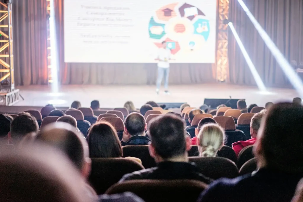 A man is standing in the stage of an auditorium while giving his speech to a crowd of conference attendees.