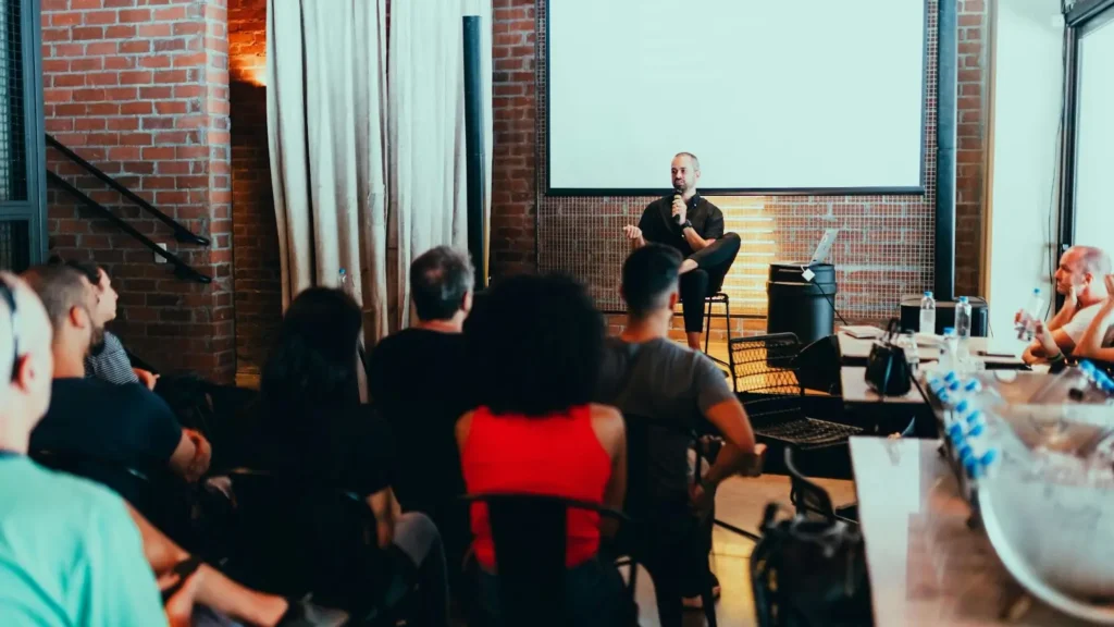 A man sitting on a chair is speaking in front of the conference attendees.